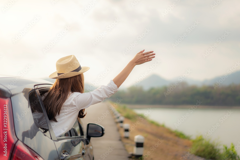 Woman at spree time relaxing and enjoying road trip. Young Asian happines female wearing hat inside compact black car with raise her hand out to the car window with mountain and lake view at sunny day