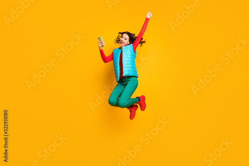Full length photo crazy ecstatic girl use smartphone read social network discount win lottery jump raise fists scream wear red blue green pants vest footwear isolated bright color background
