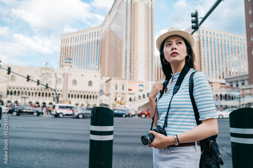 stylish girl in sunhat standing on city street on backdrop of modern skyscraper architecture. woman tourist hold camera walking in las vegas boulevard on blue sunny day. summer backpack travel