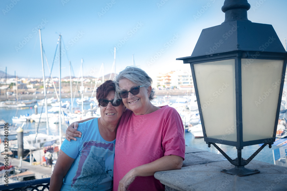 Hug between two smiling senior sisters. Couple of people, in vacation or retirement. Harbor and boats in background. Blue sky. Relaxation under the tropical sun
