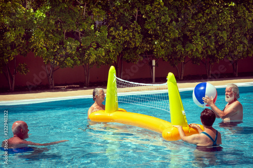 Having fun in the swimming pool with inflatable net and ball. Four people, two elderly women and men. Bright light of the sun