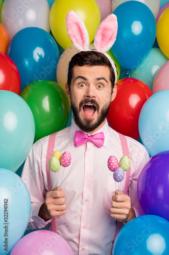 Vertical photo of funny guy colorful decor party festive mood hold painted eggs sticks open mouth wear pink shirt bow tie suspenders fluffy bunny ears on balloons creative background