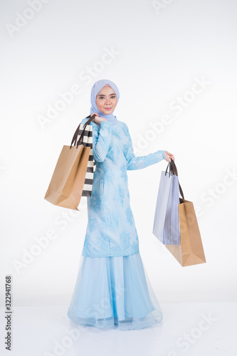 A beautiful Muslim female model in a Asian traditional dress modern kurung carrying shopping bags isolated on white background. Eidul fitri festive preparation shopping concept. Full length portrait.