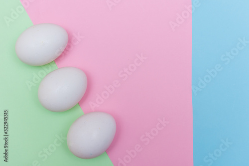 Three Easter holiday flat lay with white egg on a solid light green, blue, and pink pastel background with copy space.