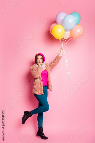 Full size vertical photo of funny pretty lady hold many colorful balloons flying up air crazy wear modern fur coat scarf beret cap trousers shoes isolated pink color background