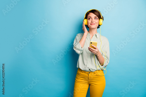 Photo of pretty lady hold telephone using modern technology listening radio earphones look empty space wear casual green shirt yellow pants isolated blue color background