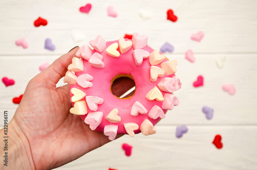 Sweet pink donut with icing and decoration with hearts on a rainbow wooden background in female hand
