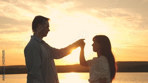 Loving man and woman dance in bright rays of sun on the background of the lake. Young couple dancing at sunset on beach. Happy guy and girl waltz in the evening in summer park.