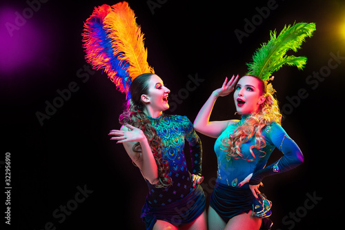 Posing. Beautiful young women in carnival, stylish masquerade costume with feathers on black background in neon light. Copyspace for ad. Holidays celebration, dancing, fashion. Festive time, party.