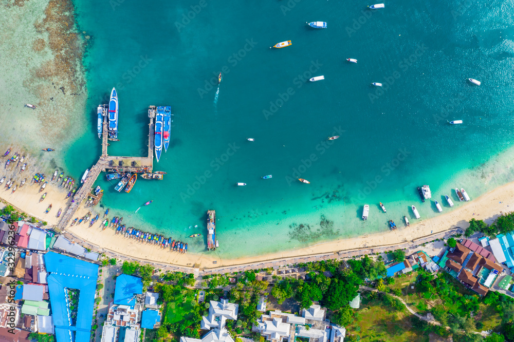 Flying above busy tropical island port filled with boats and ferries. Countless ships sail to and from bustling harbor on exotic beach. Picturesque shot of fleet of anchored longtail boats.