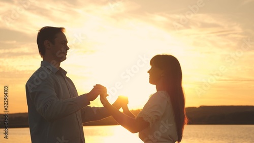 Loving man and woman dance in bright rays of sun on the background of the lake. Young couple dancing at sunset on beach. Happy guy and girl waltz in the evening in summer park.