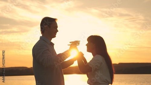 Loving man and woman dance in bright rays of sun on the background of the lake. Young couple dancing at sunset on beach. Happy guy and girl waltz in the evening in summer park.
