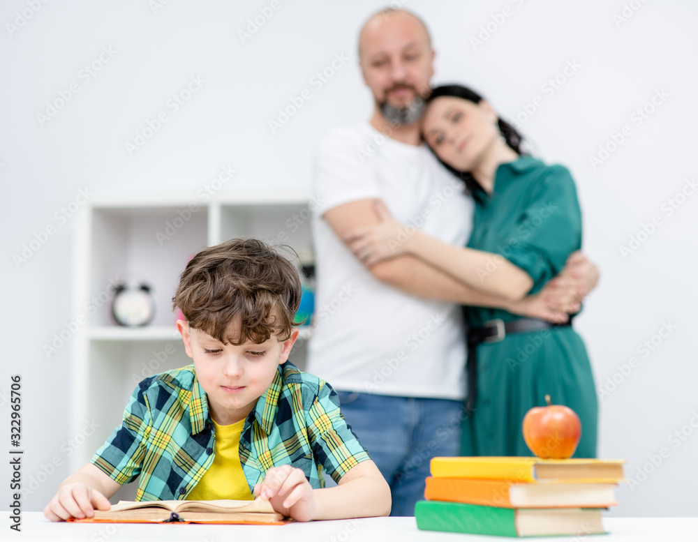 Young boy reads a book while parents embracing at looking at him at home Young boy reads a book while parents embracing at looking at him at home