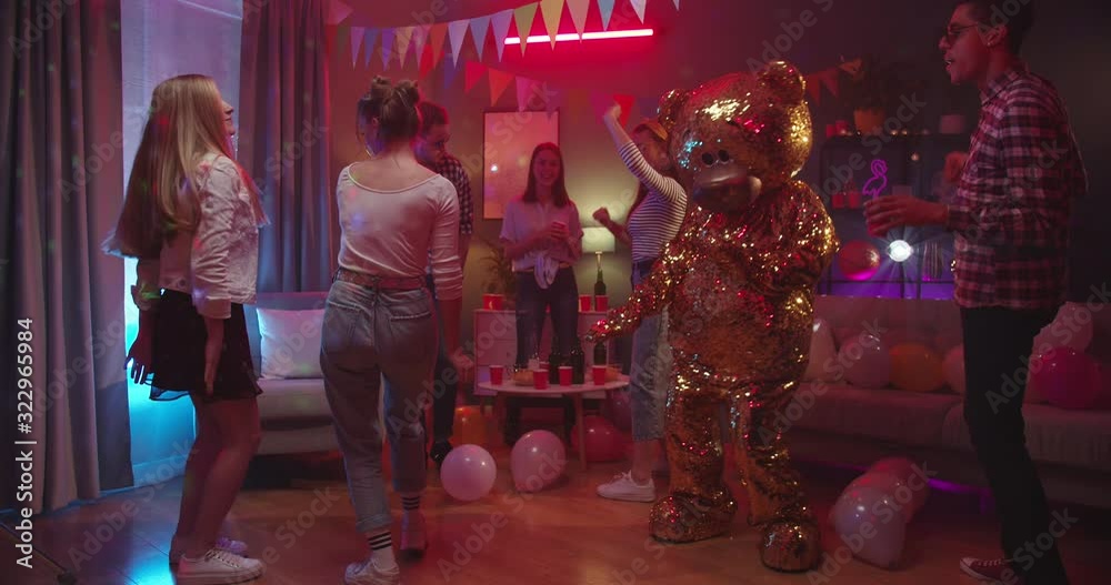 Big funny golden grown doll of teddy bear dancing at the home party among young happy guys and girls in the decorated room.