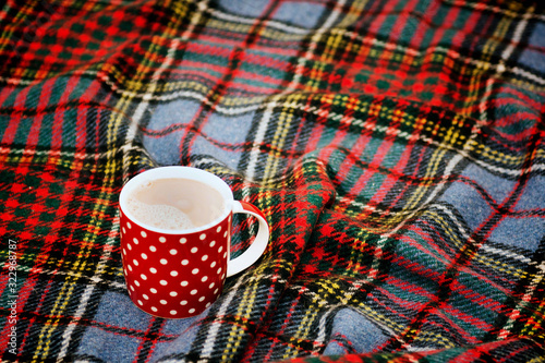 Red dotted teacup with hot chocolate on a Scottish blanket. Cozy home concept with red porcelain mug. A Cup of festive hot chocolate. Traditional homemade Christmas advertising cocoa. New year's