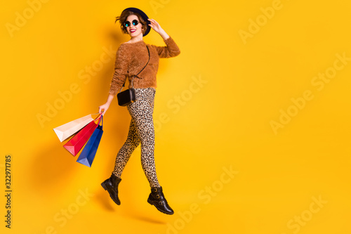 Full length photo of funky lady jump high sales shopping carry packs black friday wear fluffy pullover leopard pants shoes retro cap clutch specs isolated yellow color background