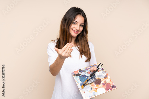 Young artist man holding a palette isolated on beige background inviting to come