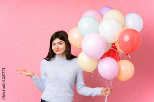 Young Ukrainian teenager girl holding lots of balloons over isolated pink background having doubts with confuse face expression