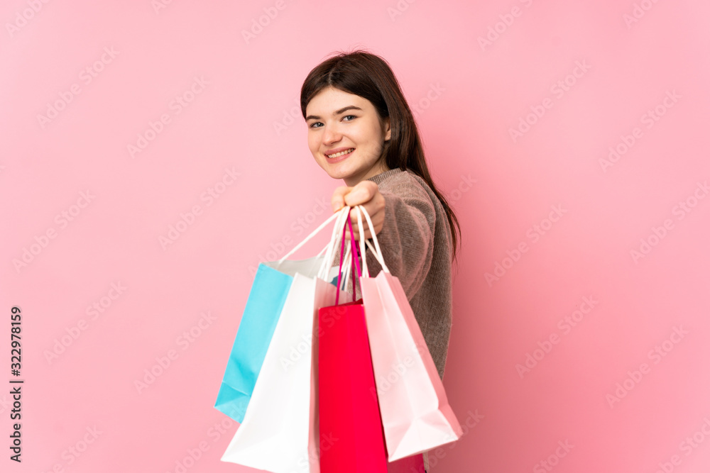 Young Ukrainian teenager girl over isolated pink background holding shopping bags and giving them to someone