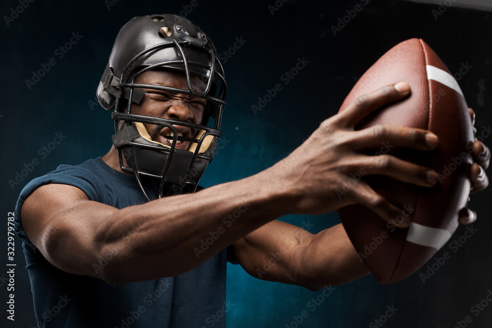 portrait photo of dark-skinned young man on a dark background wearing a t-shirt, rugby helmet and holds rugby ball in his arms he has anger on his face