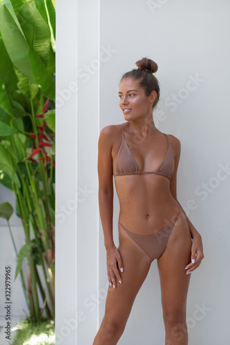 Slim tanned girl in beige bikini posing near white wall of tropical villa
