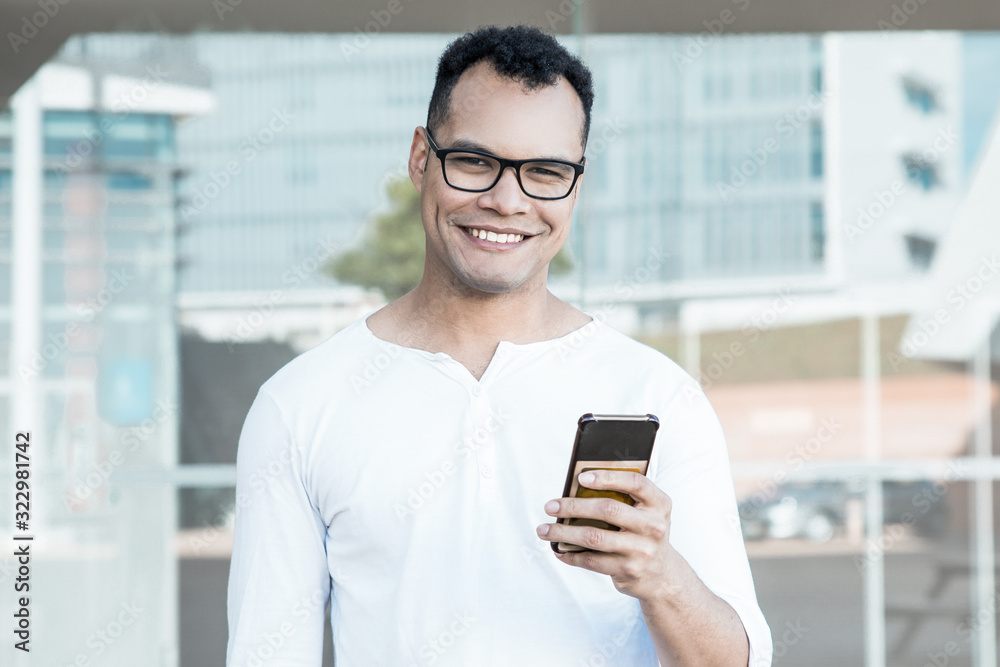 Medium shot of young mixed-race man in spectacles and white T-shirt standing at office building, holding phone in hands, looking at camera, smiling. Lifestyle concept Medium shot of young mixed-race man in spectacles and white T-shirt standing at office building, holding phone in hands, looking at camera, smiling. Lifestyle concept