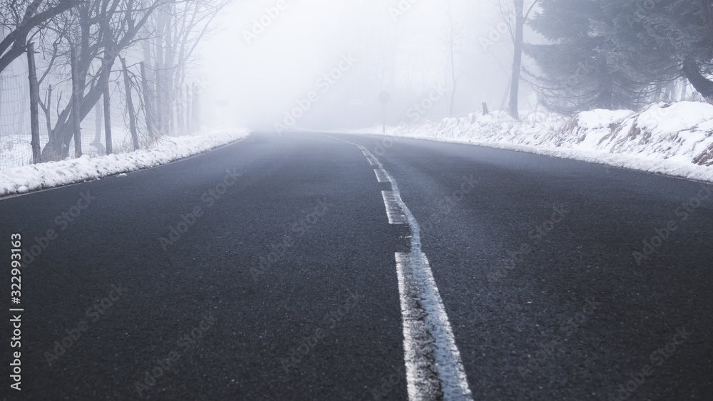 An asphalt road in a rural setting leading into thick fog in the distance on a snowy winters day. An asphalt road in a rural setting leading into thick fog in the distance on a snowy winters day.