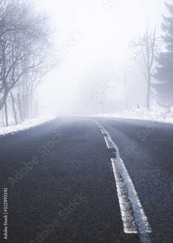 An asphalt road in a rural setting leading into thick fog in the distance on a snowy winters day.