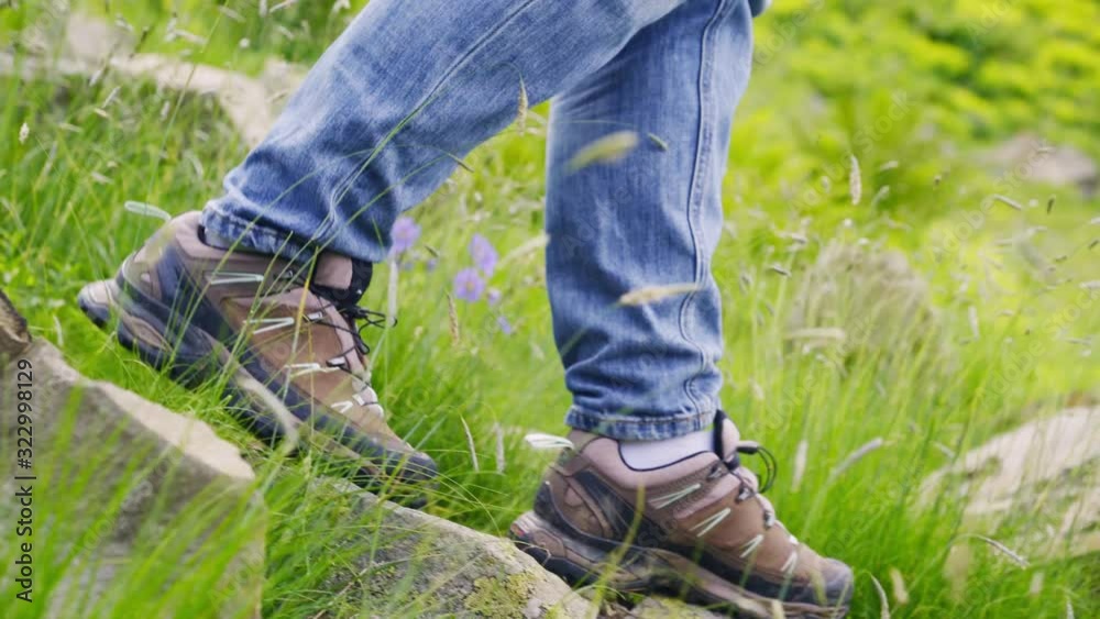 Woman backpacker walking down on rocky path in sunny day. Following faceless female goes uphill along flowering alpine meadow, detail shot feet in trekking boots stepping from stone to stone Woman backpacker walking down on rocky path in sunny day. Following faceless female goes uphill along flowering alpine meadow, detail shot feet in trekking boots stepping from stone to stone