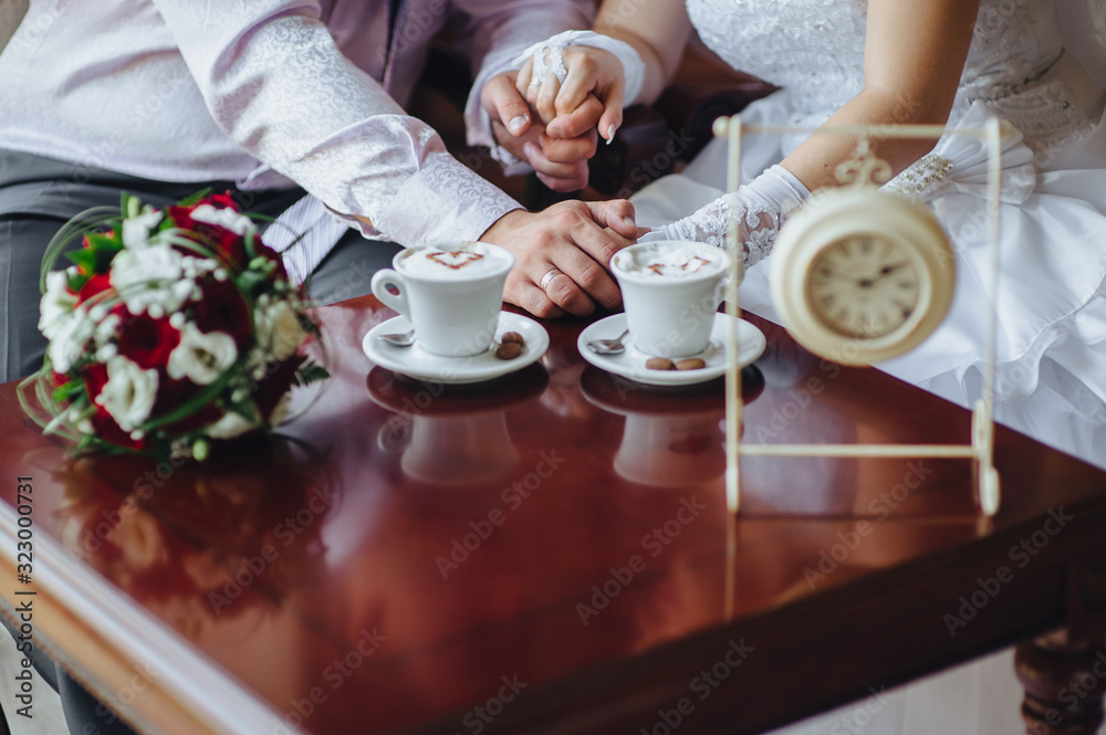 The bride and groom tenderly hold hands, sitting in a cafe with cups of coffee with foam in the form of a heart, and a bouquet of roses and eustomas. Photography, concept. The bride and groom tenderly hold hands, sitting in a cafe with cups of coffee with foam in the form of a heart, and a bouquet of roses and eustomas. Photography, concept.