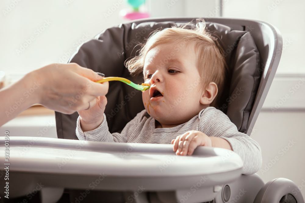 Beautiful baby in the child dining chair eat healthy food. Mother's hands. Child eat vegetable puree. Lifestyle. Sunny day. Beautiful baby in the child dining chair eat healthy food. Mother's hands. Child eat vegetable puree. Lifestyle. Sunny day.