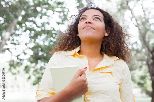 Dreamy young woman holding book in city park. Woman looking upwards and standing with blurred trees in background. Summer and education concept. Front view.