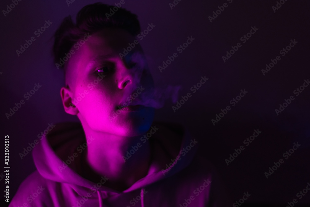 A young guy smokes cigarettes in a neon light. The problem of drug trafficking among adolescents. Teenage boy soars vape in the studio