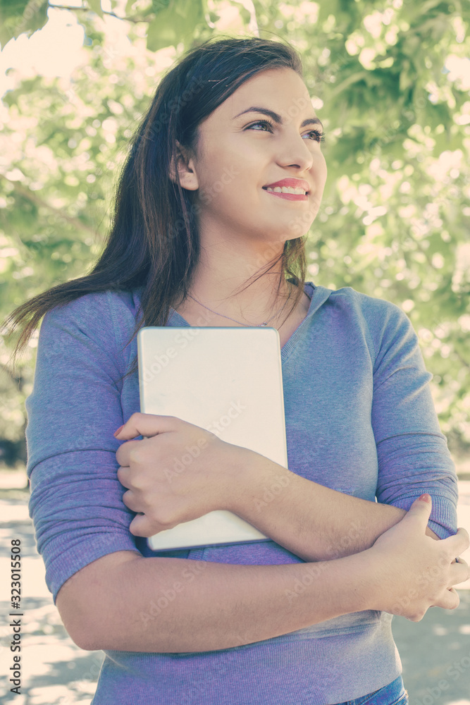 Dreamy pretty young lady holding tablet computer in park. Beautiful woman wearing hoodie, standing and looking away with green trees in background. Leisure and nature concept. Front view. Dreamy pretty young lady holding tablet computer in park. Beautiful woman wearing hoodie, standing and looking away with green trees in background. Leisure and nature concept. Front view.