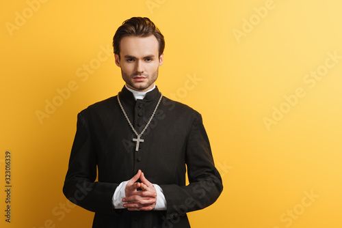 serious catholic priest standing with clenched hands while looking at camera isolated on yellow photo