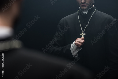 cropped view of catholic priest touching cross on his necklace near own reflection isolated on black