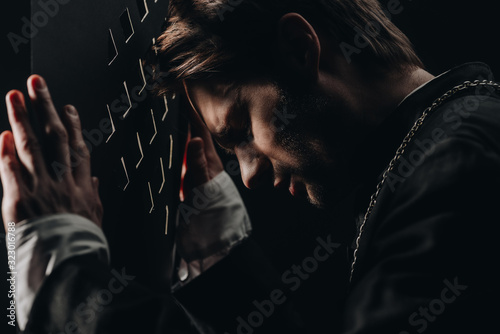 young tense catholic priest with closed eyes leaning on confessional grille in dark with rays of light photo