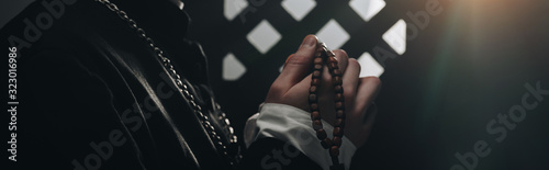 Partial view of catholic priest holding wooden rosary beads near confessional grille in dark with rays of light, panoramic shot