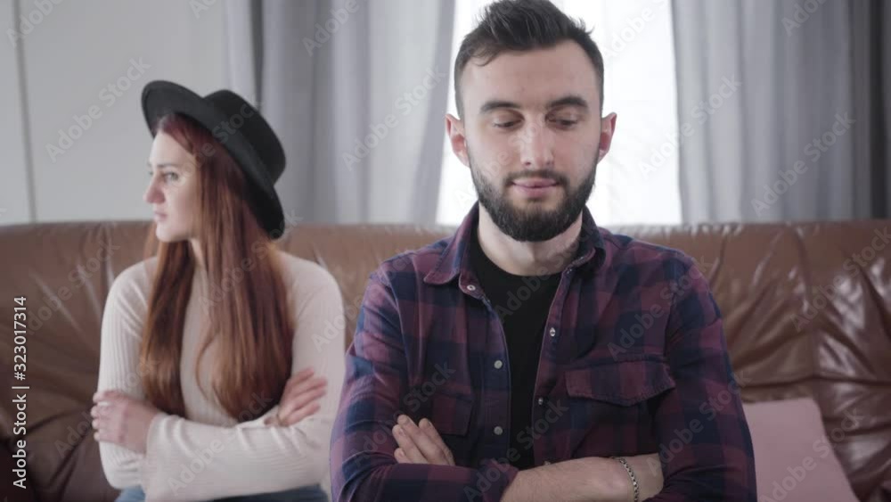 Portrait of young handsome Caucasian man with brown eyes and brunette hair sitting with crossed hands as his sad wife or girlfriend crying at the background. Concept of relationship problems, breakup Portrait of young handsome Caucasian man with brown eyes and brunette hair sitting with crossed hands as his sad wife or girlfriend crying at the background. Concept of relationship problems, breakup