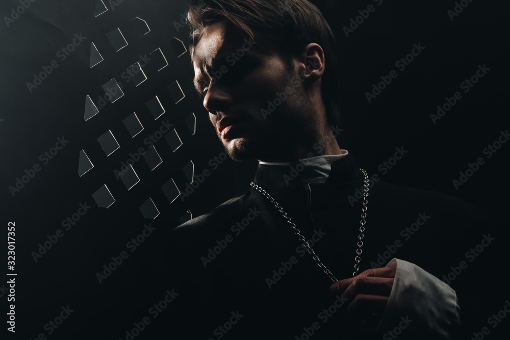 young serious catholic priest touching cross on his necklace in dark near confessional grille - obrazy, fototapety, plakaty 