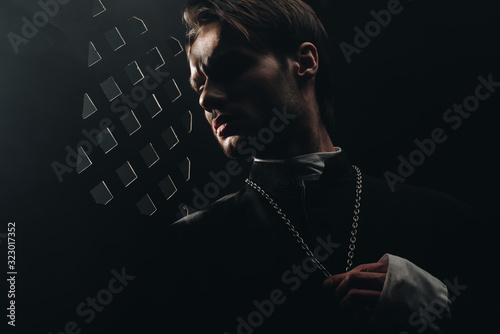 young serious catholic priest touching cross on his necklace in dark near confessional grille photo