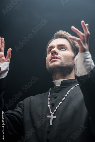 low angle view of young serious catholic priest praying with raised hands isolated on black