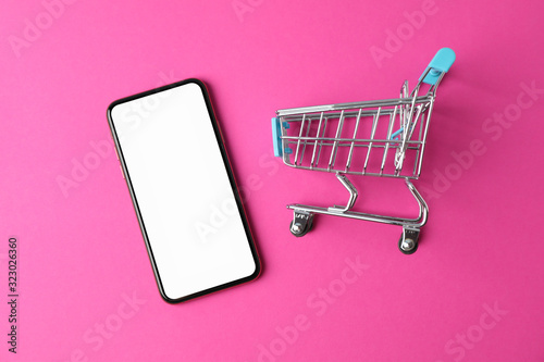 Phone with empty screen and shopping cart on pink background, top view