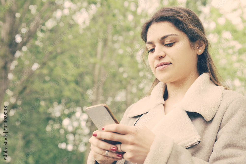 Content pretty young woman using smartphone in park. Lady wearing jacket, texting on gadget and standing with green trees in background. Communication and nature concept. Content pretty young woman using smartphone in park. Lady wearing jacket, texting on gadget and standing with green trees in background. Communication and nature concept.