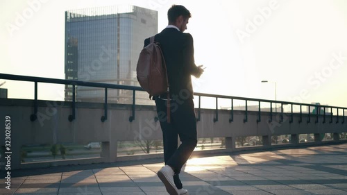 A young businessman walks around the city at a sunrise, drinks coffee and dials a message on his smartphone. Back view. Heavy traffic road in the background. Business