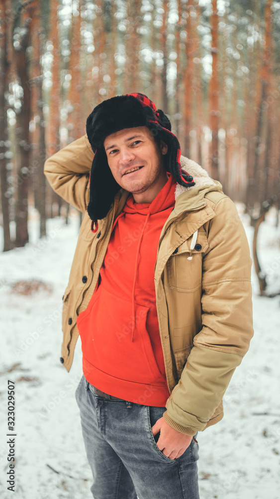 Handsome man standing in forest in winter season. Brutal young male wearing beige jacket over red hoodie and earflap hat posing in snowy forest. Handsome man standing in forest in winter season. Brutal young male wearing beige jacket over red hoodie and earflap hat posing in snowy forest.