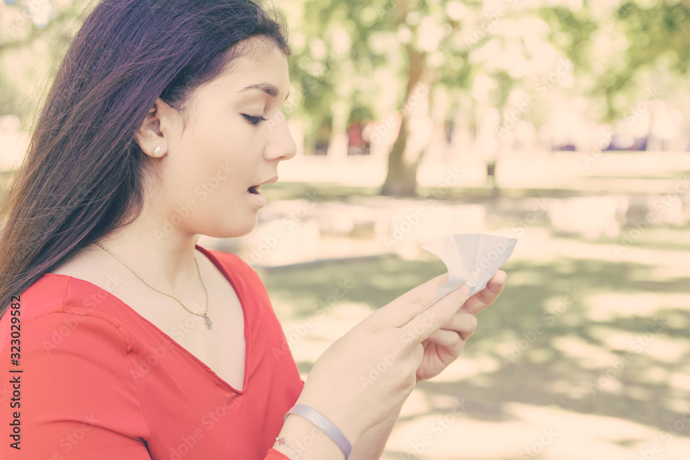 Pretty young woman holding tissue and sneezing in park. Beautiful lady wearing red blouse and standing with green lawn and trees in background. Cold and nature concept. Side view. Pretty young woman holding tissue and sneezing in park. Beautiful lady wearing red blouse and standing with green lawn and trees in background. Cold and nature concept. Side view.