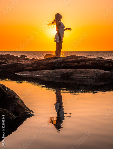 Lifestyle, silhouette of a young blonde in a white dress on the coast in a sunset moving her hair, reflected in the water
