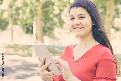 Happy pretty young woman browsing on tablet in park. Beautiful lady wearing red blouse and looking at camera with green trees in background. Technology and nature concept.