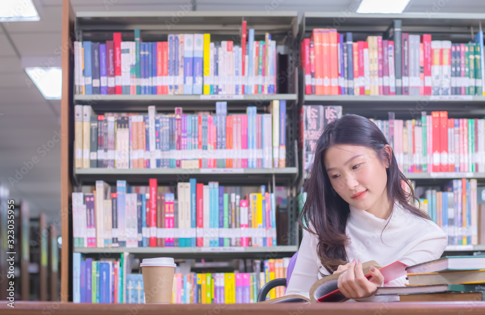 Young Asian smile female student portrait sitting and concentrate studying or reading textbook in university library for exams or test with book shelves background. Education and learning concept Young Asian smile female student portrait sitting and concentrate studying or reading textbook in university library for exams or test with book shelves background. Education and learning concept
